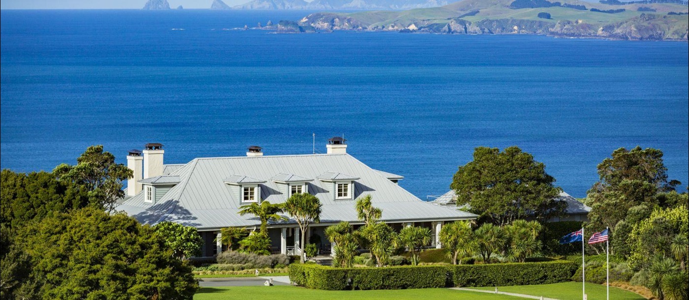 Exterior shot of Kauri Cliffs Lodge in New Zealand with green lawns in front and the Pacific Ocean and mountains in the background