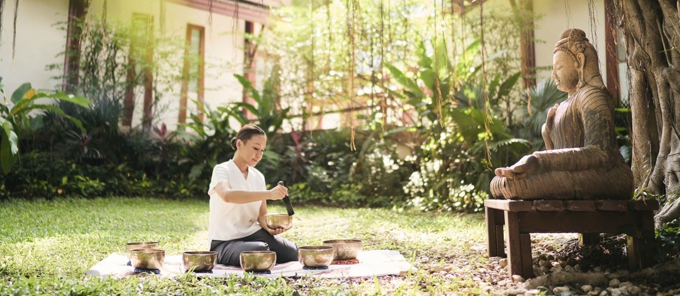 Lady performing Tibetan Sound healing bowls in a garden in front of a buddha statue at Luxury resort Chiva Som