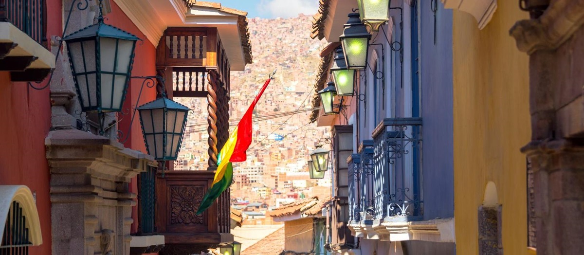 A street in La Paz with red, blue and yellow painted houses that have balconies and lights overlooking the street. A Bolivian flag is flying from a wooden balcony on the left.