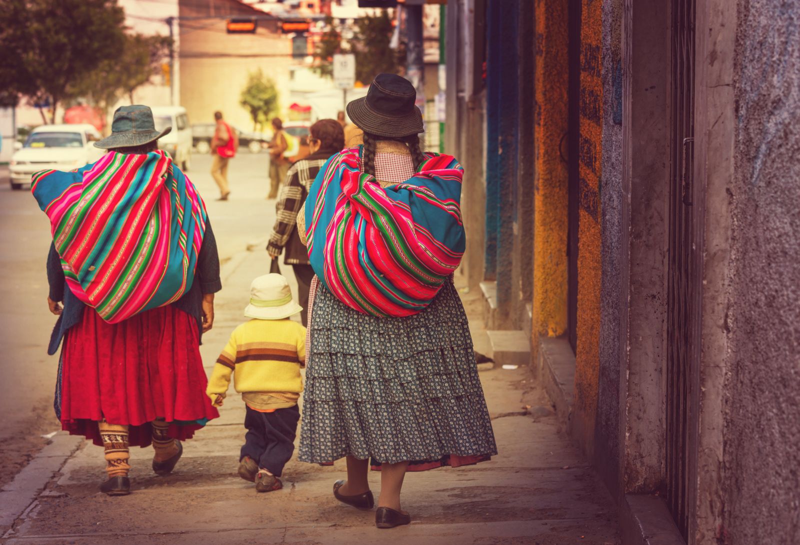 Two women carrying belongings in coloured blankets on their backs walking down the street in Boliva with a small child in between them