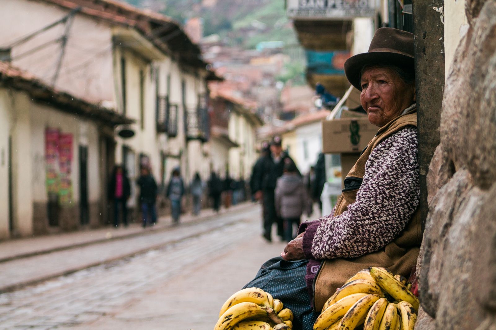 A woman dressed in a sweater and brown waistcoat and wearing a hat sitting in a street in Bolivia with bunches of bananas