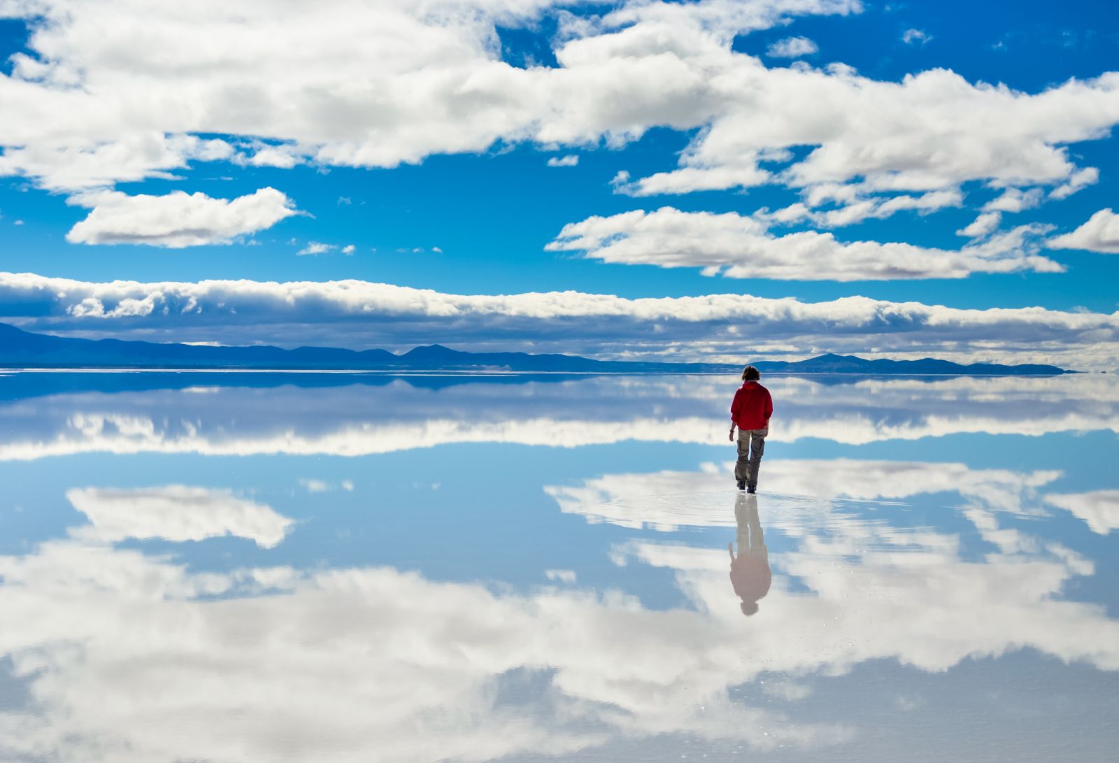 A lady in a red jacket walking across the salt flats in Bolivia on a sunny day with mountains in the background 