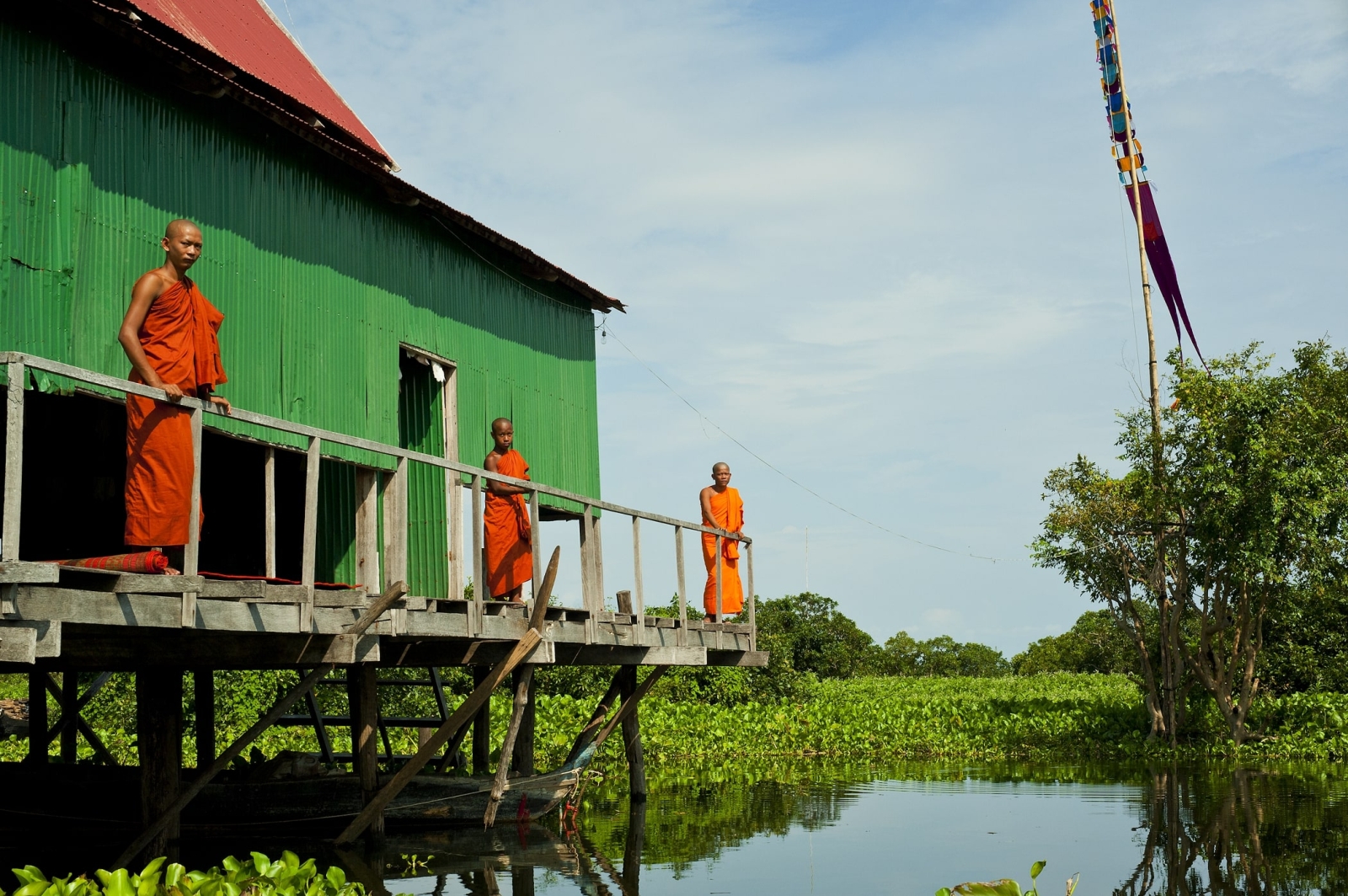 Three monks stand by a stilted green house on the Mekong