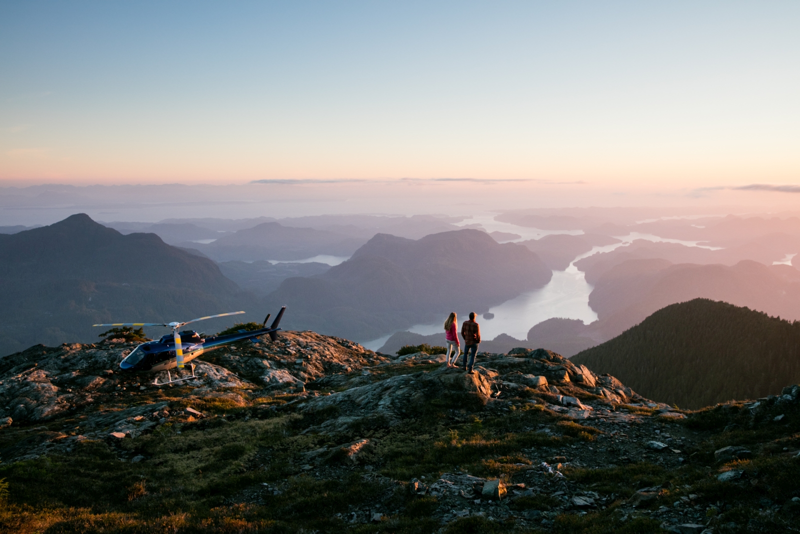 Two guests on top of a mountain with a helicopter, enjoying the incredible views over the surrounding mountains and bays at luxury hotel Nimmo Bay in Canada