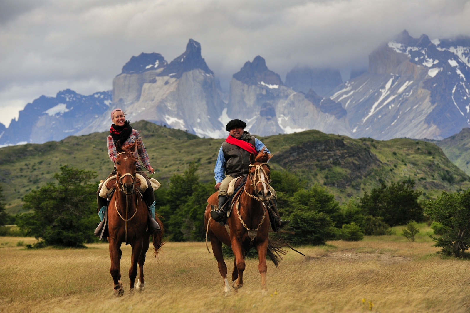 Two people riding horses with mountain range backdrop 