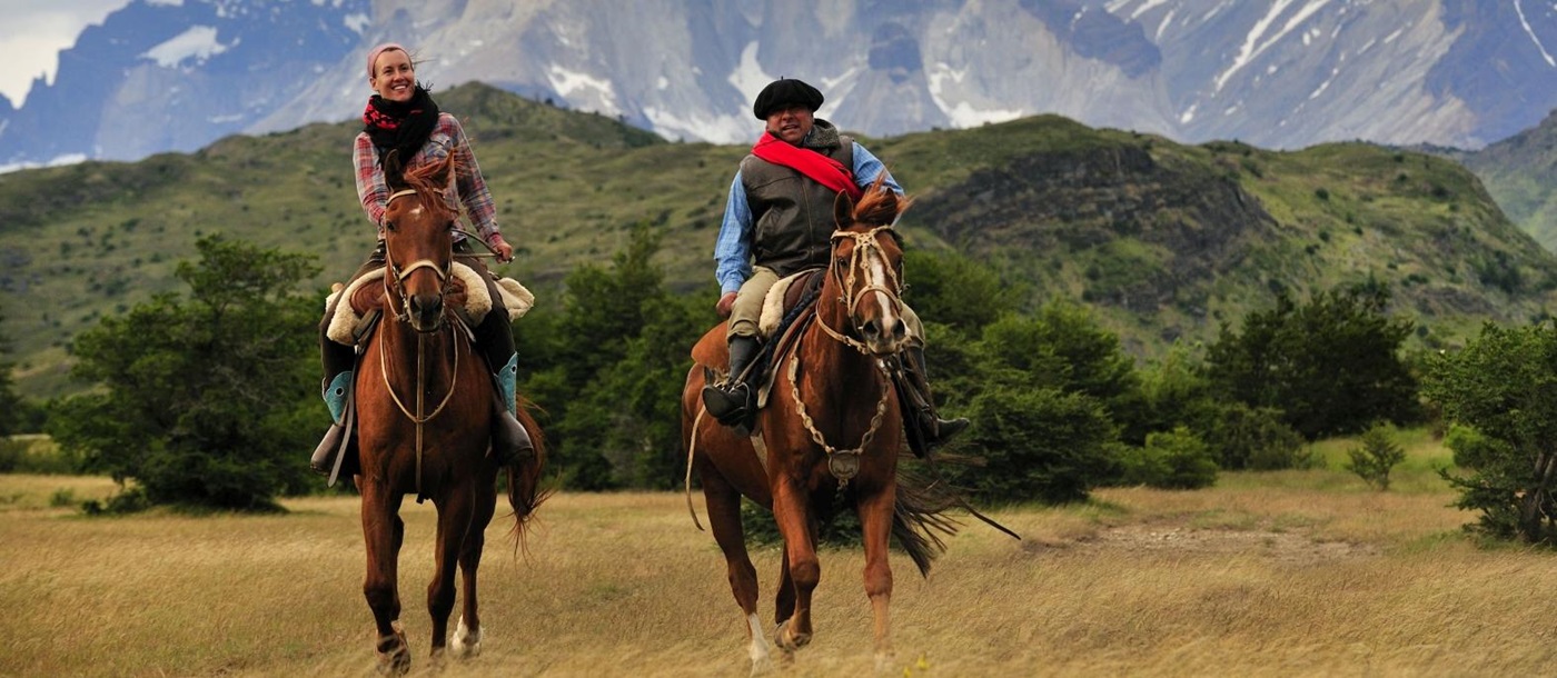 Two people riding horses with mountain range backdrop 