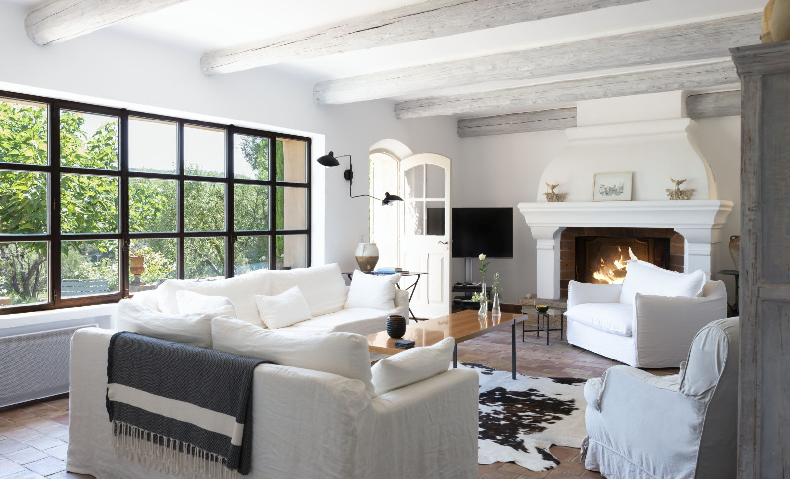 Living room with sofas, armchairs, tables, fireplace, ceiling beams, lamps & window at La Cadiere on the Cote d’Azur, France