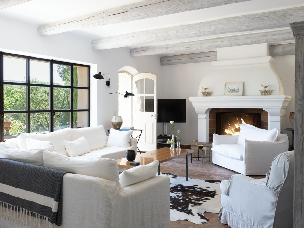 Living room with sofas, armchairs, tables, fireplace, ceiling beams, lamps & window at La Cadiere on the Cote d’Azur, France