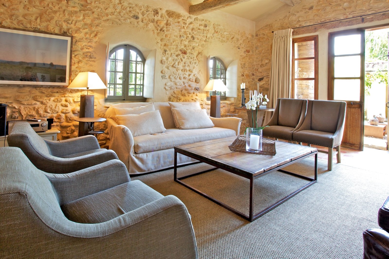 Living room with armchairs, sofa, tables, lamps and French doors to patio at Le Pont Romain in Provence, France