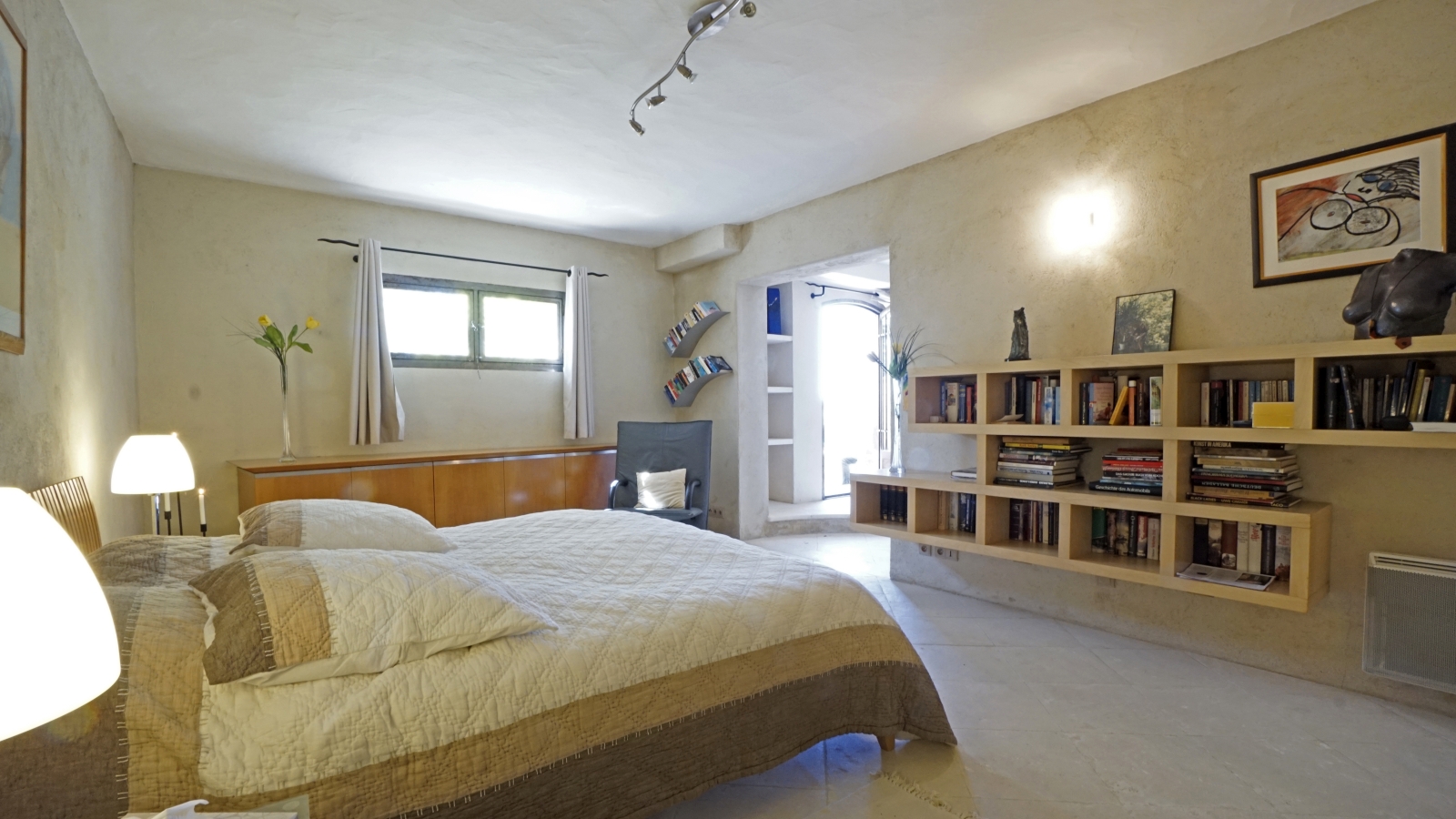 Bedroom with double bed, lamps, bookshelves, artwork and armchair at Les Rochers in Provence, France