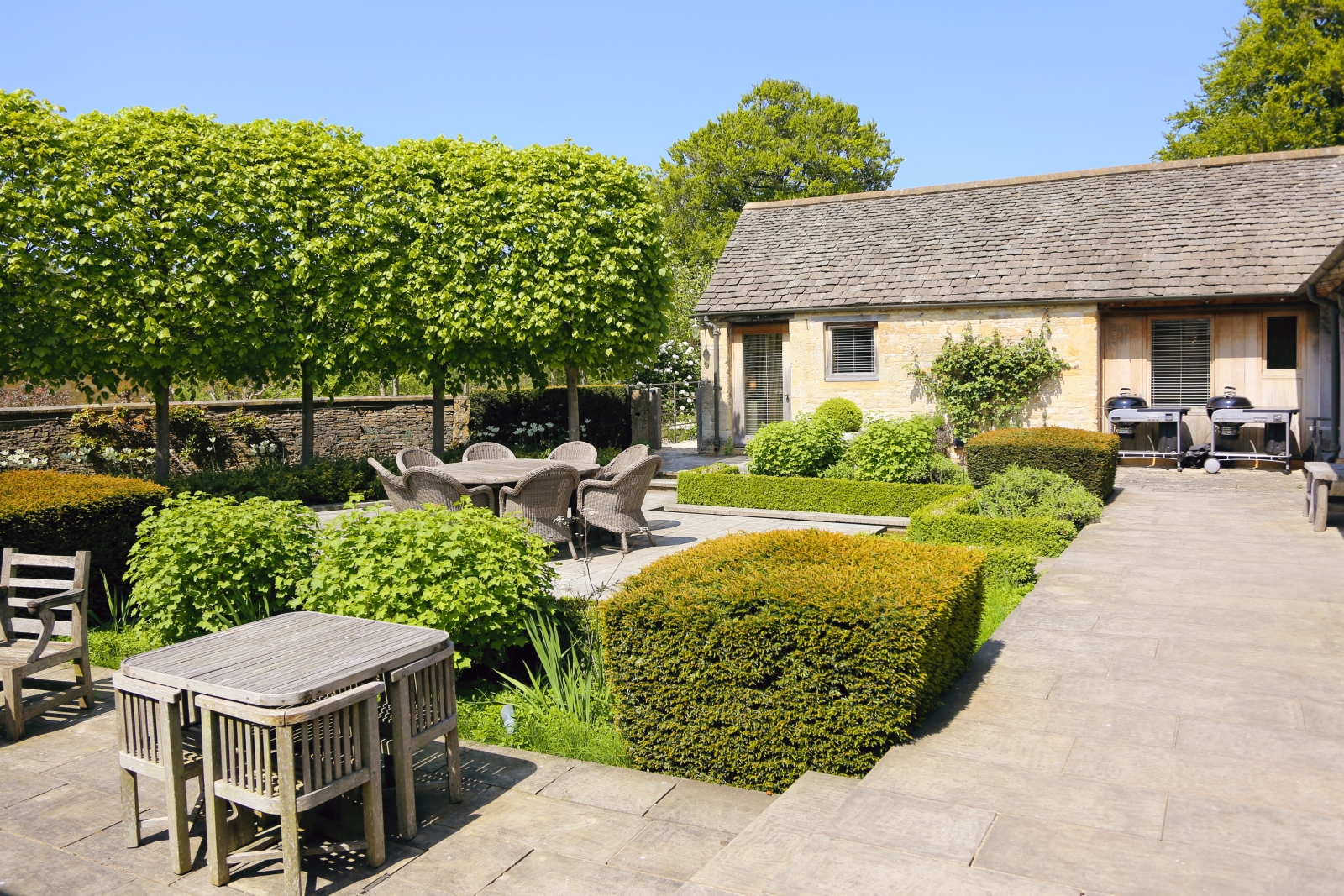 Cotswold stone back of villa with tables, chairs, barbeques, hedges & trees at Temple Guiting Barn in the Cotswolds, England