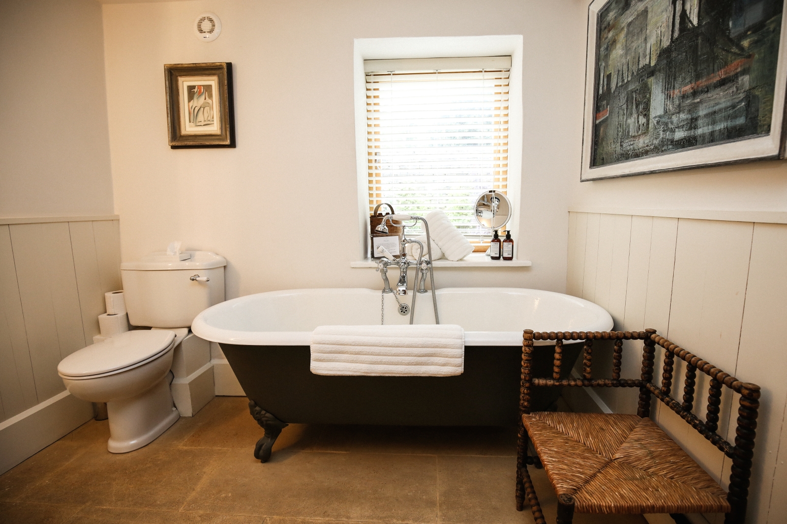 Bathroom with chair, bath, toilet, paintings and window at The Estate at Temple Guiting in the Cotswolds, England