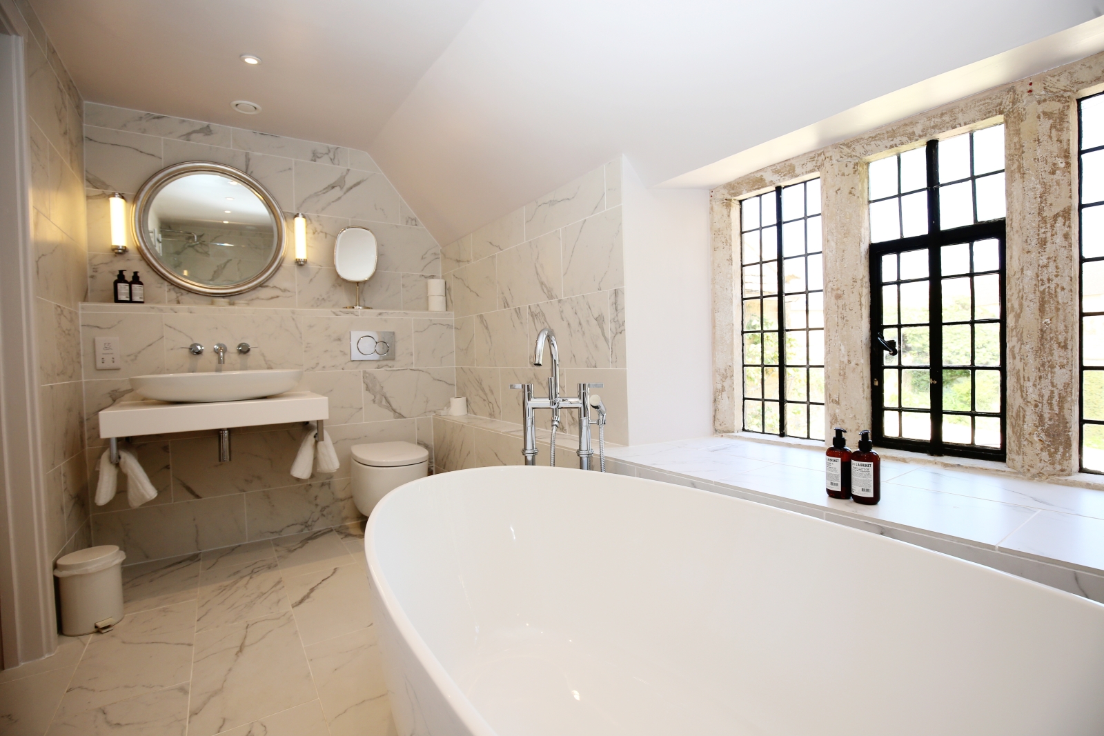 Bathroom with bath, toilet, sink, mirror, windows and marble walls at The Estate at Temple Guiting in the Cotswolds, England
