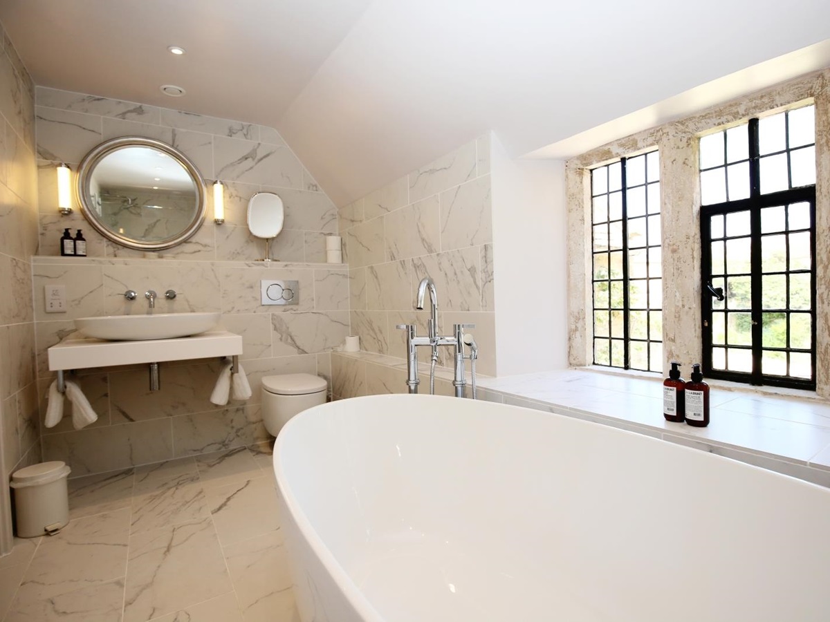 Bathroom with bath, toilet, sink, mirror and large window at Temple Guiting Manor in the Cotswolds, England
