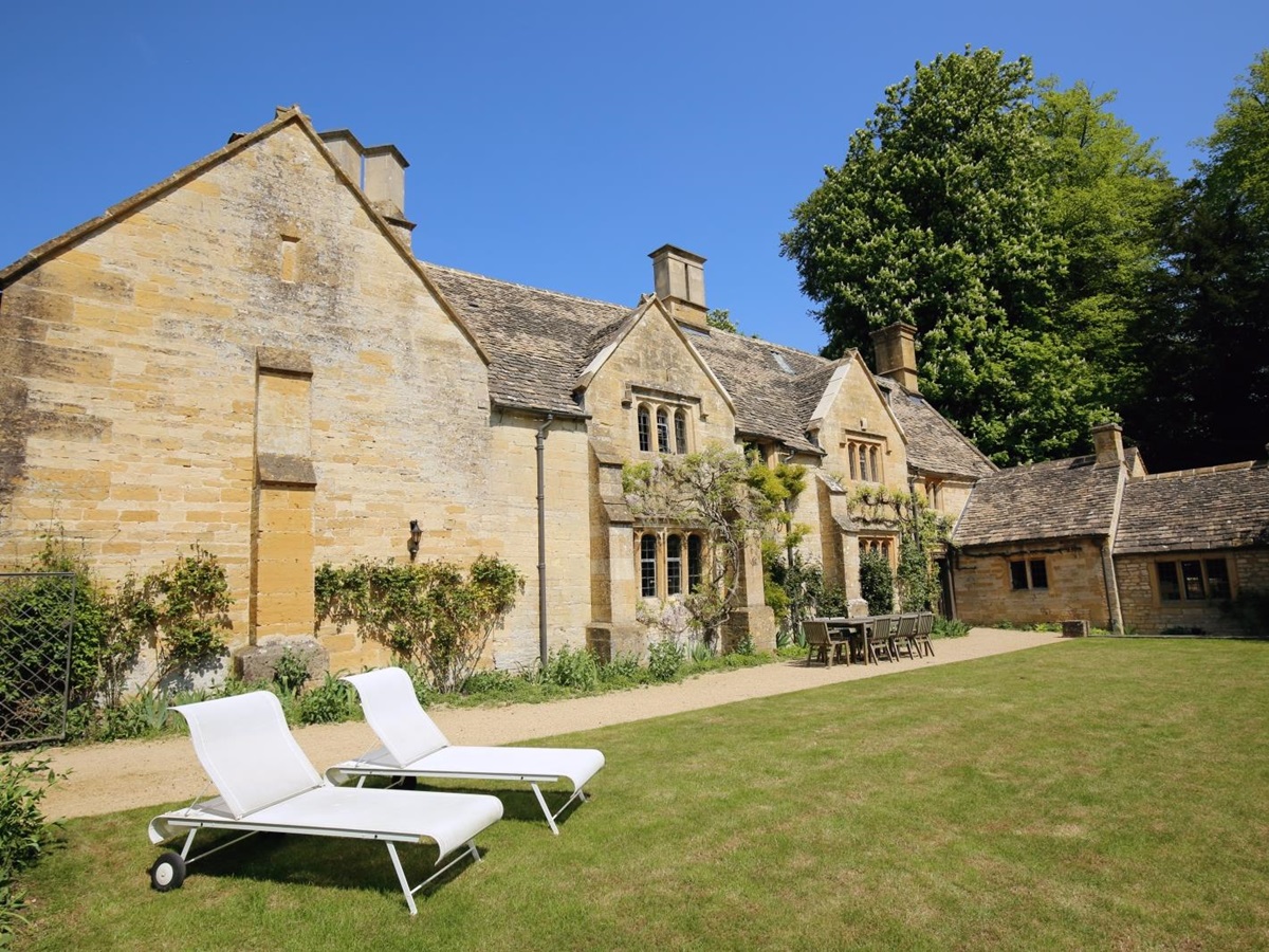 Cotswold stone back of house with garden, table, chairs and sun loungers at Temple Guiting Manor in the Cotswolds, England
