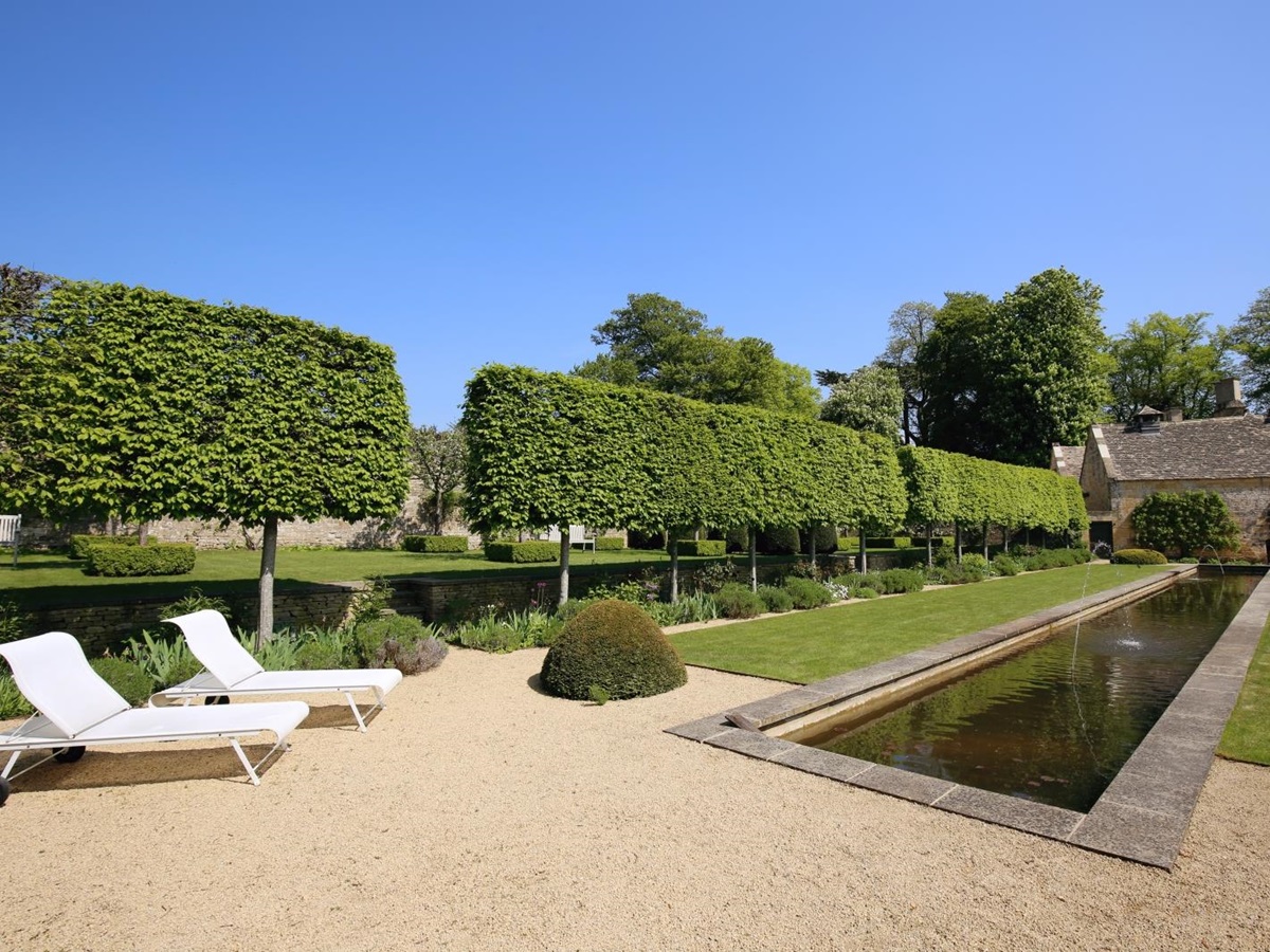 Garden with sun loungers, trees, hedges, flowers and lake with fountains at Temple Guiting Manor in the Cotswolds, England