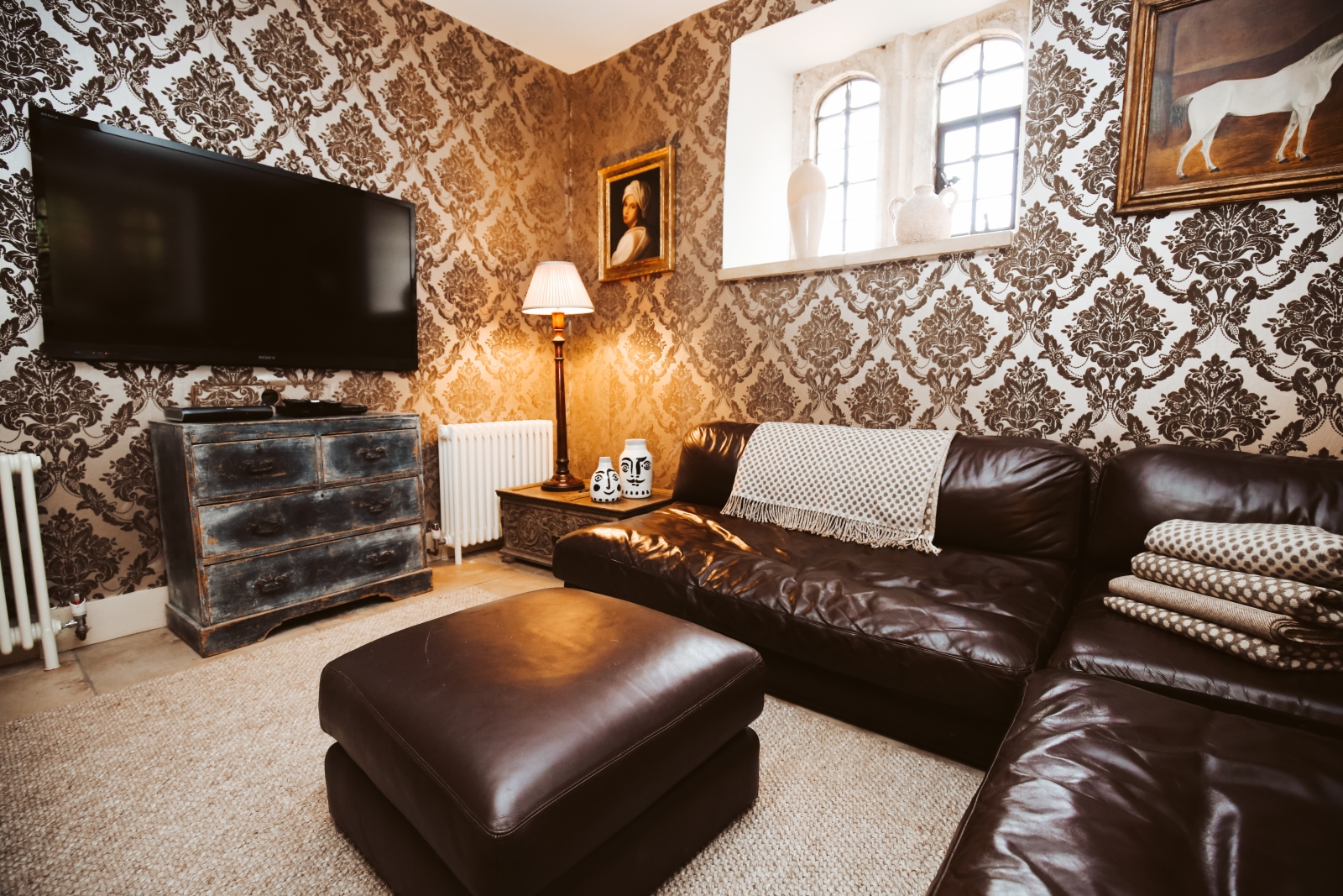 Lounge with long leather sofa, footstool, drawers, TV and paintings at Temple Guiting Manor in the Cotswolds, England