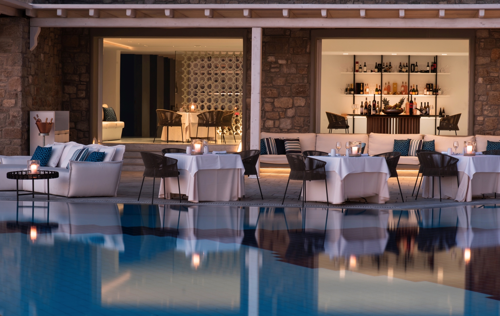 View over the pool towards Nama Restaurant, with dining tables set along the illuminated water at luxury resort Mykonos Grand, Greece