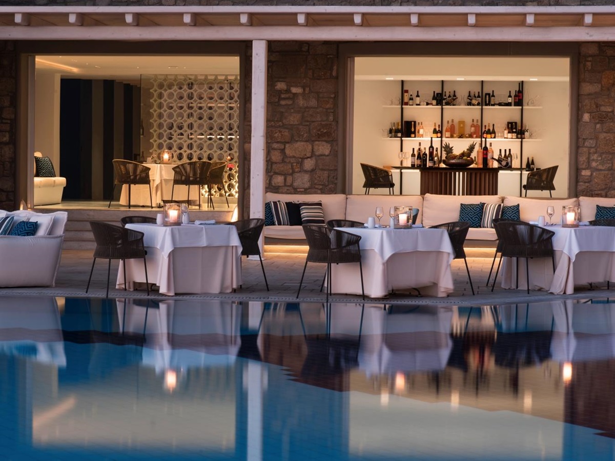 View over the pool towards Nama Restaurant, with dining tables set along the illuminated water at luxury resort Mykonos Grand, Greece