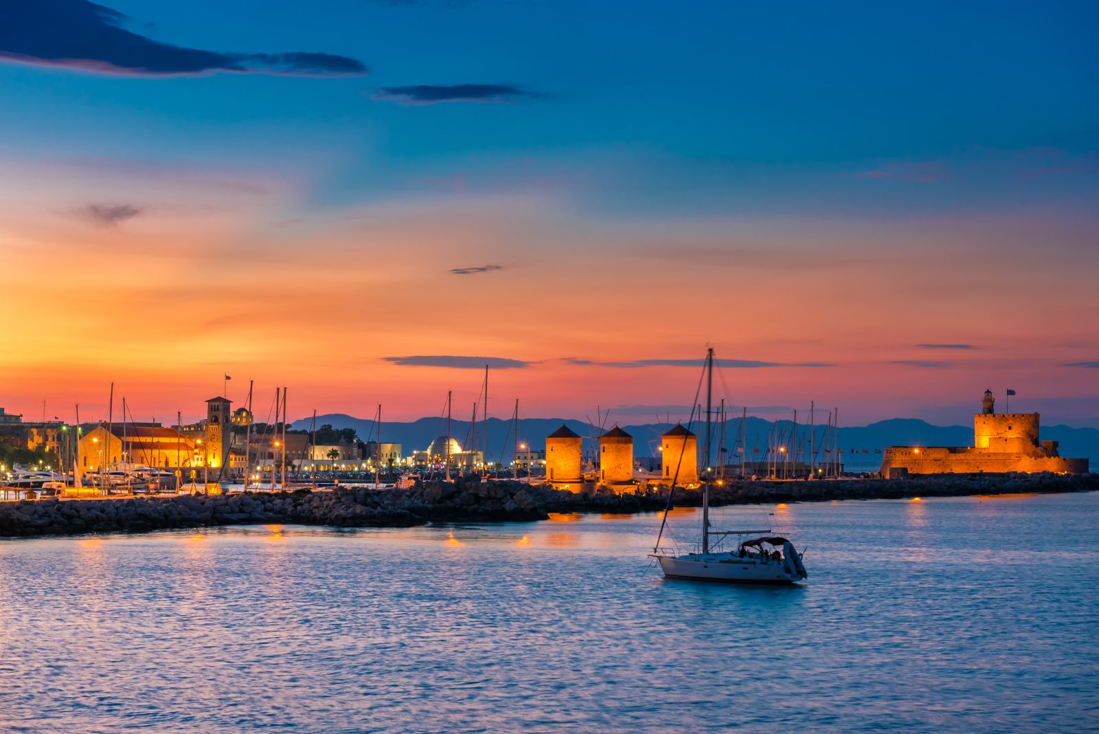 A yacht in harbour in Rhodes at sunset with illuminated windmills