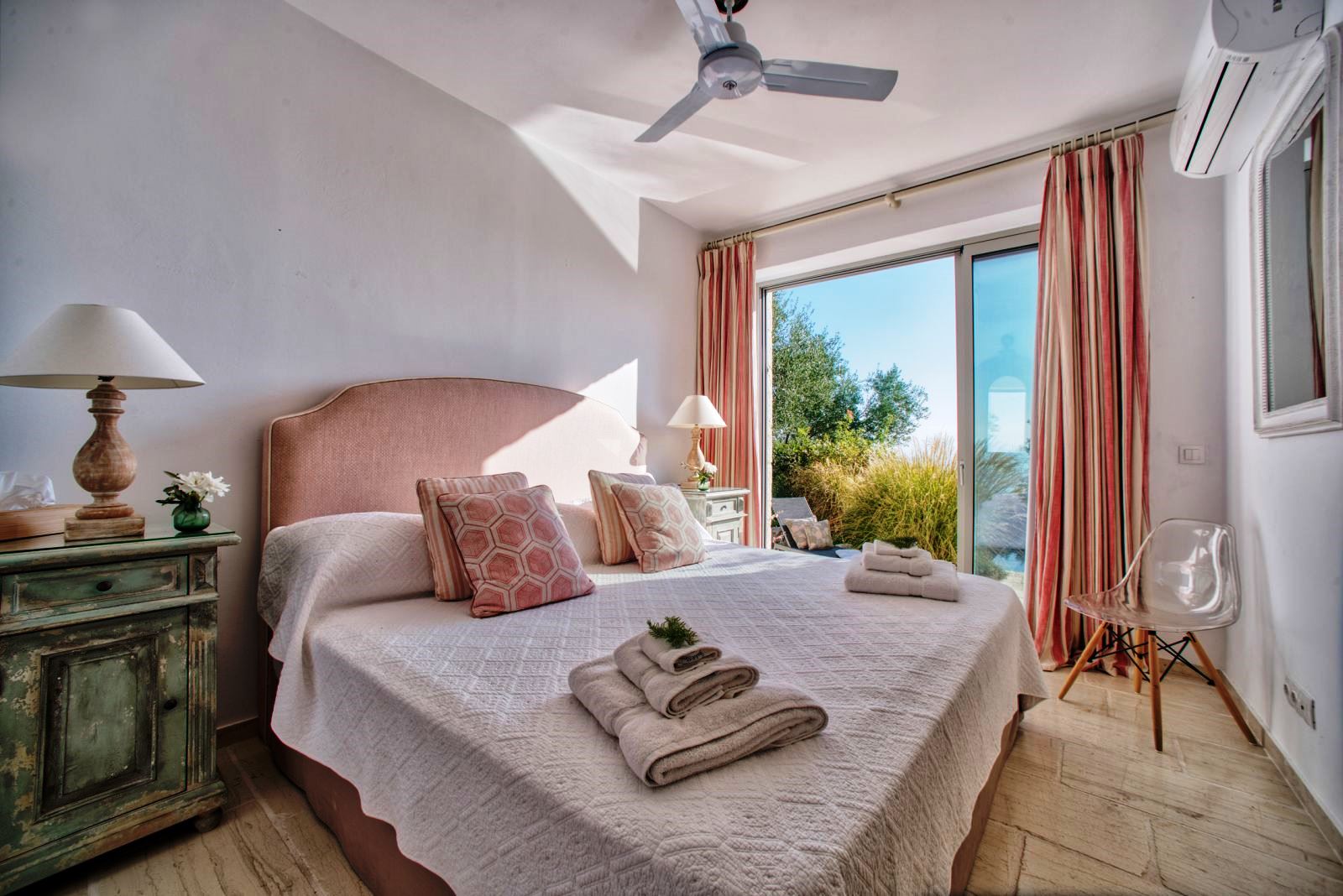 double bed with pink headboard on wooden floor and sliding glass doors onto sun terrace at villa apraos in corfu, Greece