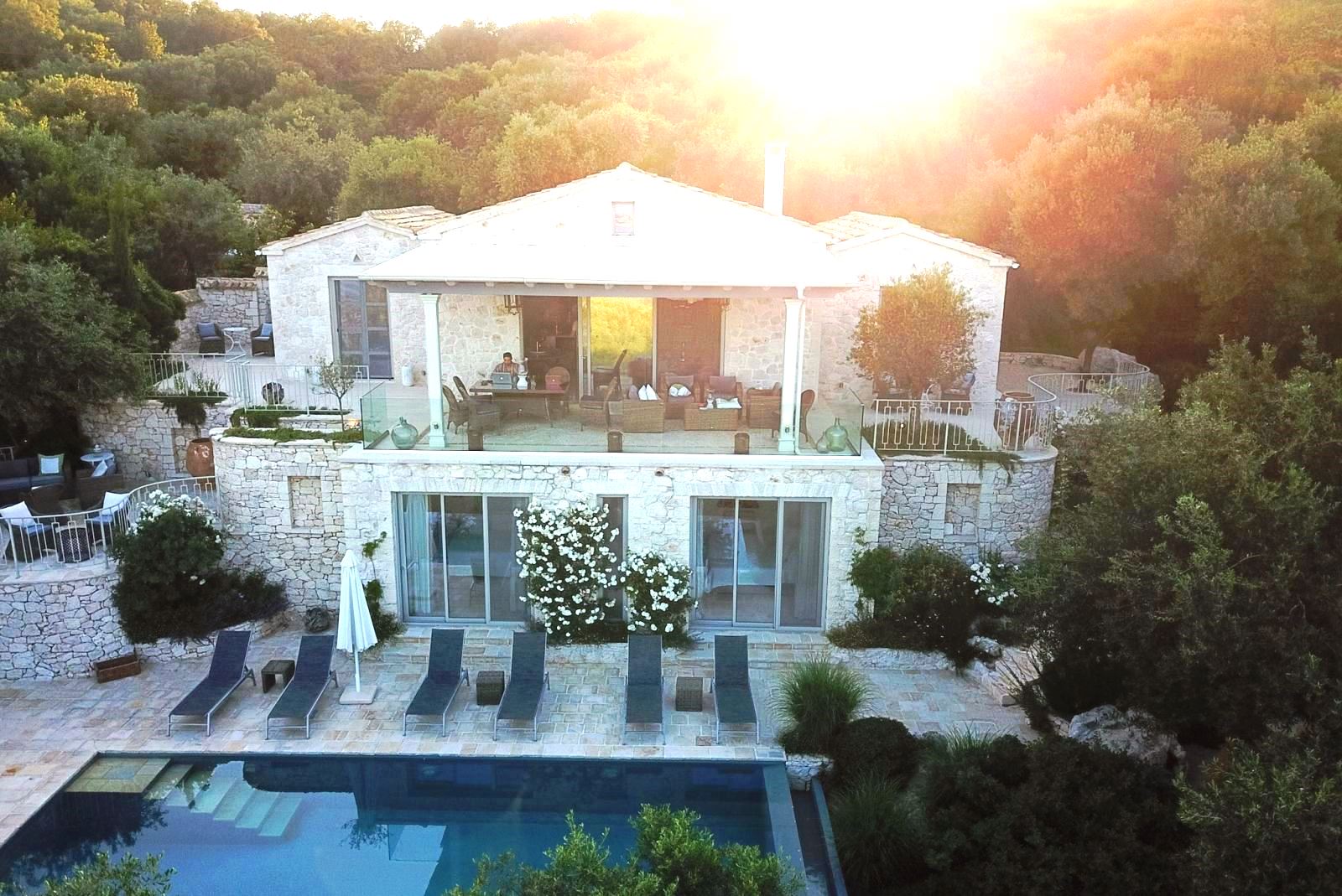 exterior view of villa apraos in corfu, Greece overlooking the pool and 6 blue sun beds with the sun behind the trees