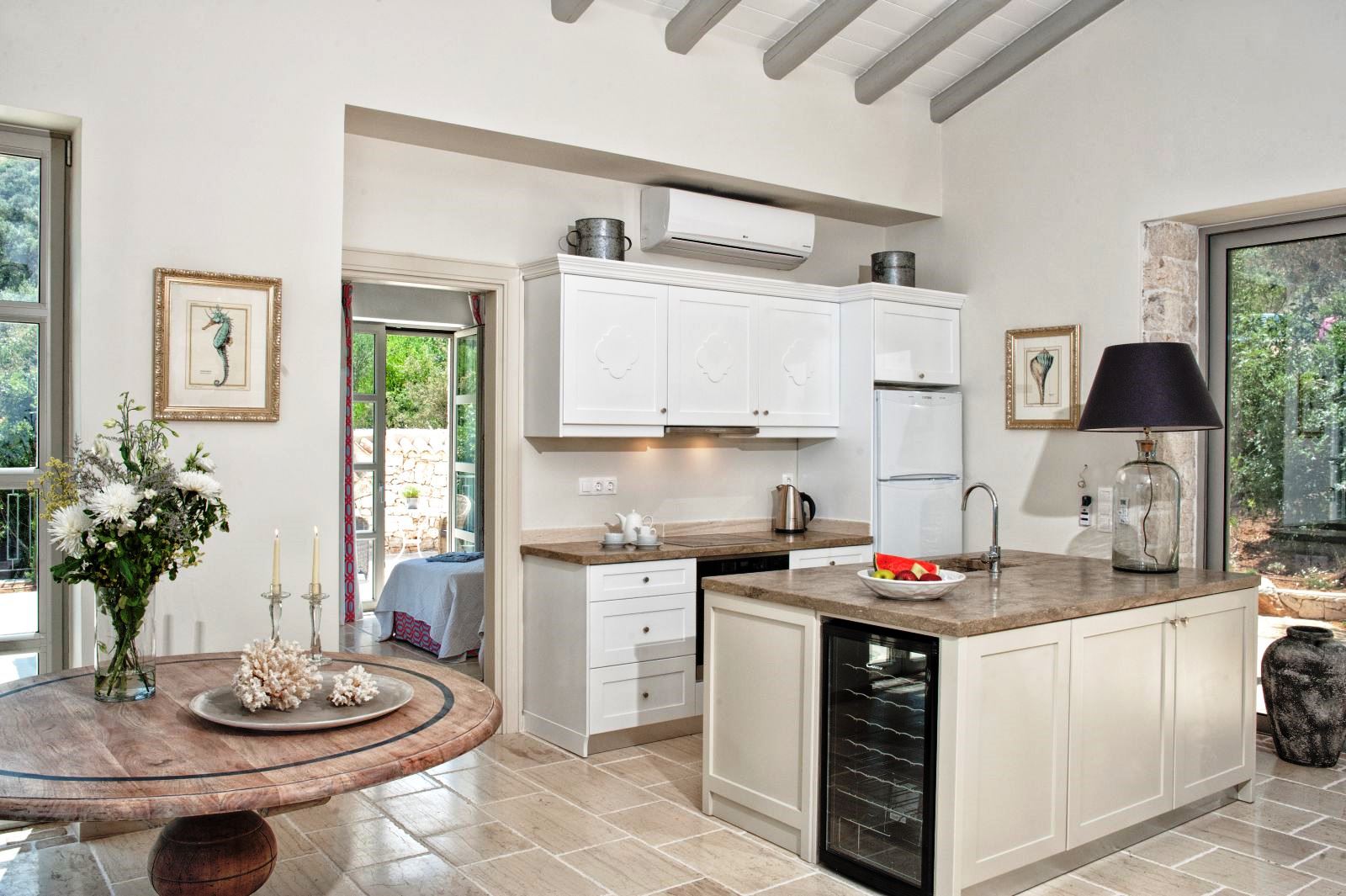 kitchen with white units, stone tiled floor and round wooden table with vase of flowers at villa apraos in corfu, Greece