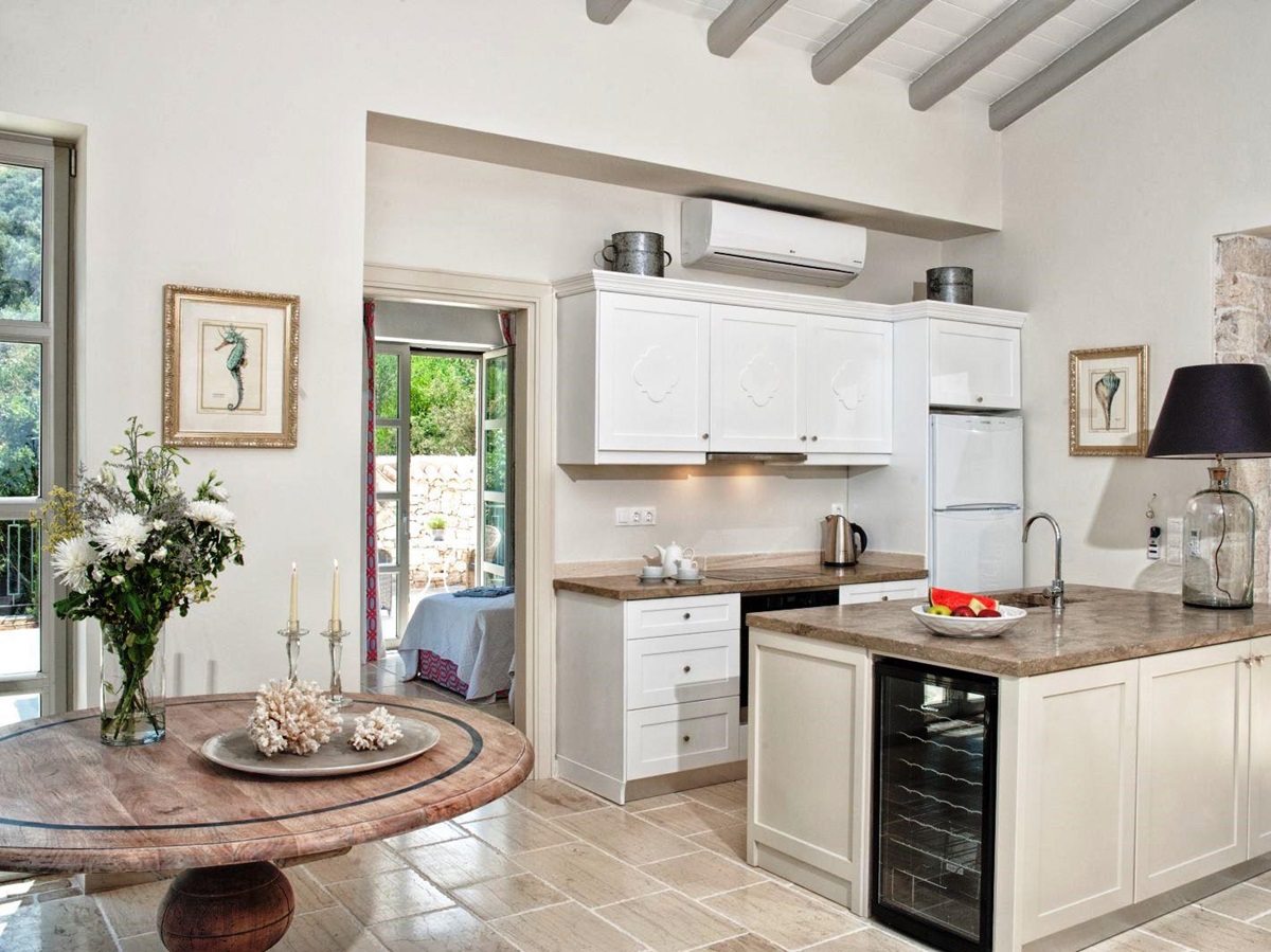 kitchen with white units, stone tiled floor and round wooden table with vase of flowers at villa apraos in corfu, Greece
