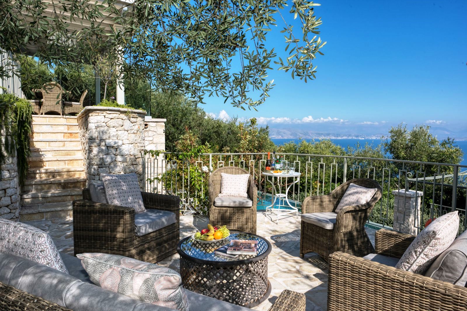 Sun terrace with wicker chairs and round table and stone steps up to dining terrace at villa apraos in corfu, Greece