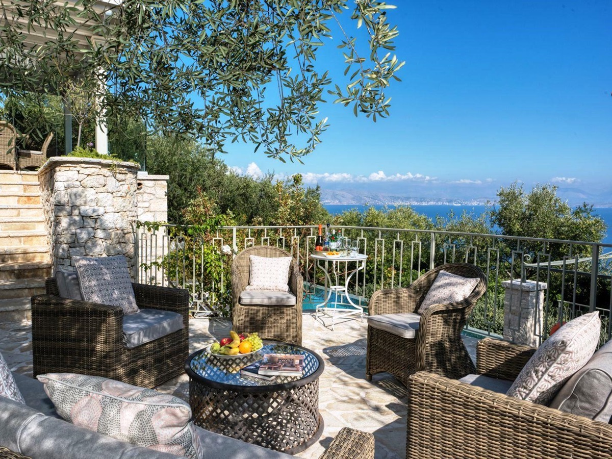 Sun terrace with wicker chairs and round table and stone steps up to dining terrace at villa apraos in corfu, Greece