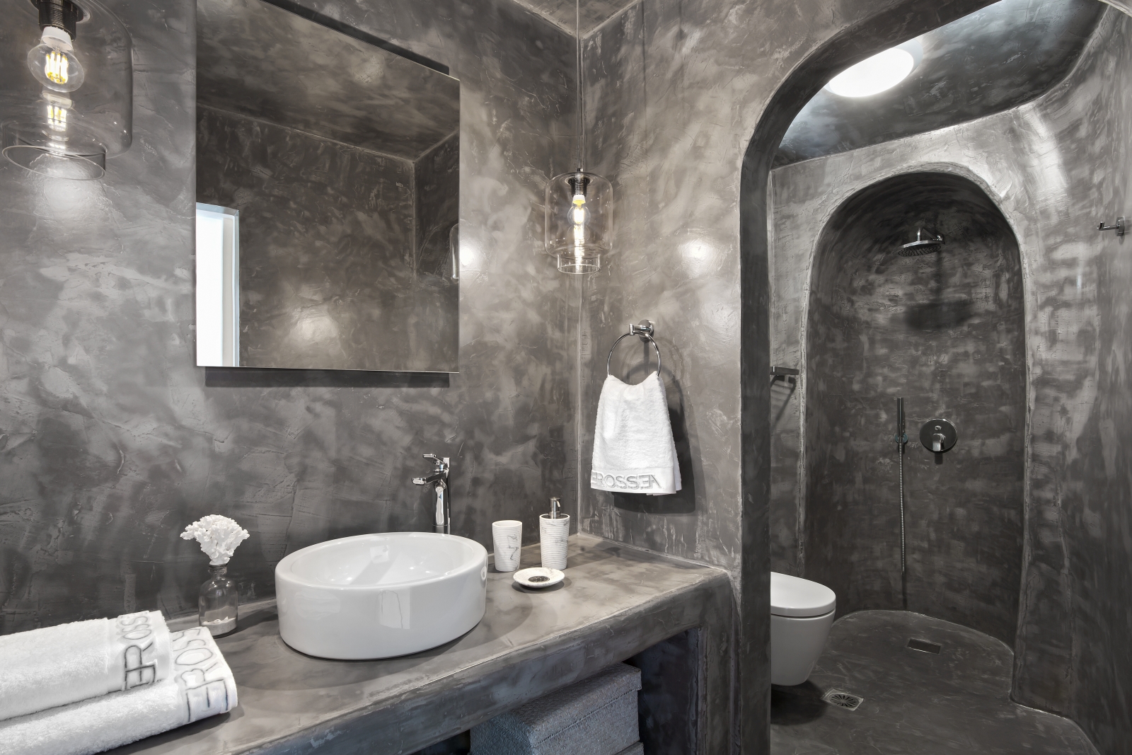 Bathroom with grey walls, sink, hanging lights, mirror, toilet and shower at Erossea on Santorini, Greece