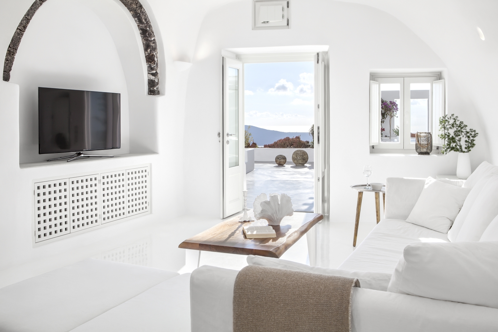 Living room with sofa, coffee table, TV, plant and doors to balcony at Erossea on Santorini, Greece