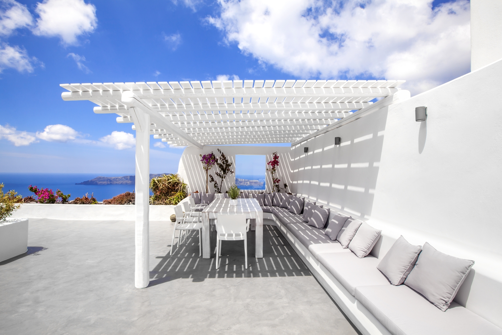 Outdoor dining area with long sofa, table, chairs, flowers and sea view at Erossea on Santorini, Greece