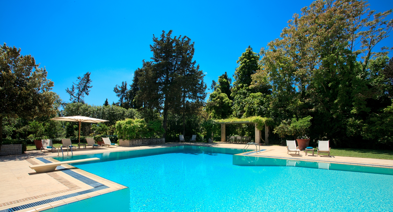 Large pool with diving board and patio area with sun loungers, umbrellas and trees at the Kanoni Estate on Corfu, Greece