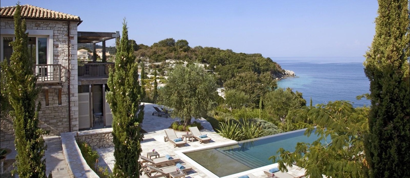 View of villa with patio, sun loungers, towels, pool, trees and sea view at the Odysseus Estate on Corfu, Greece