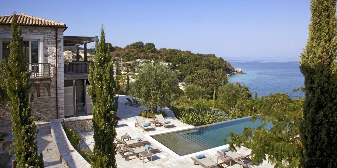 View of villa with patio, sun loungers, towels, pool, trees and sea view at the Odysseus Estate on Corfu, Greece