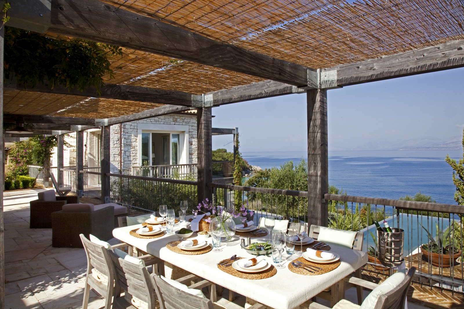 Covered balcony with table, chairs, crockery, comfy armchairs & view of pool and sea at the Odysseus Estate on Corfu, Greece