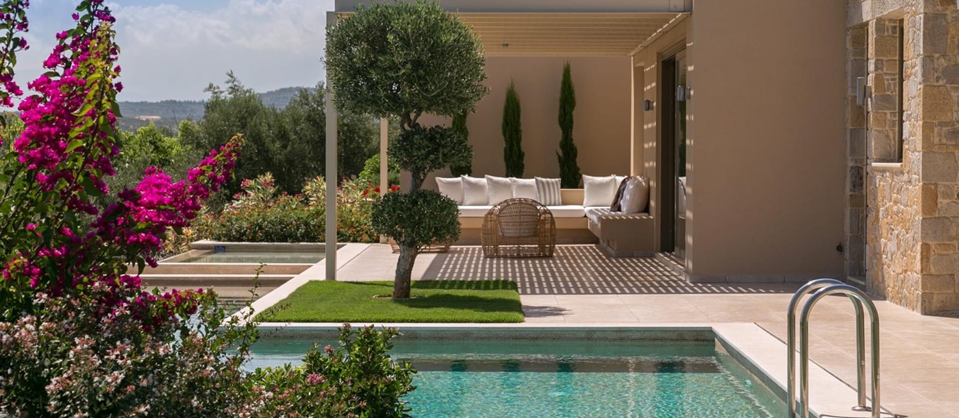 View of pool and outdoor lounge area with comfy chairs and sofa and flowers in foreground at Villa Ianira on Crete, Greece