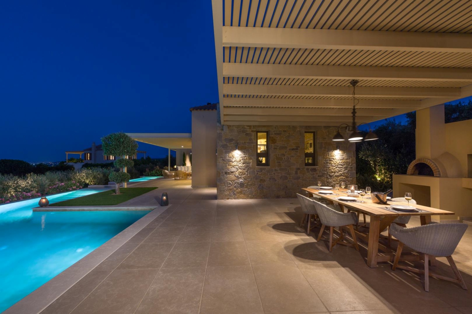 Covered dining area at night with table, chairs, wood oven, pool and large patio area at Villa Ianira on Crete, Greece