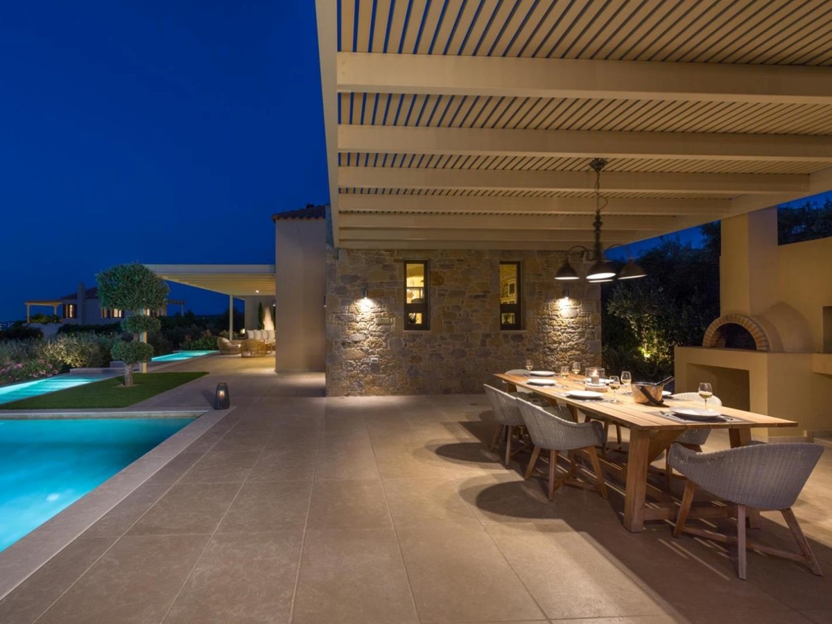 Covered dining area at night with table, chairs, wood oven, pool and large patio area at Villa Ianira on Crete, Greece