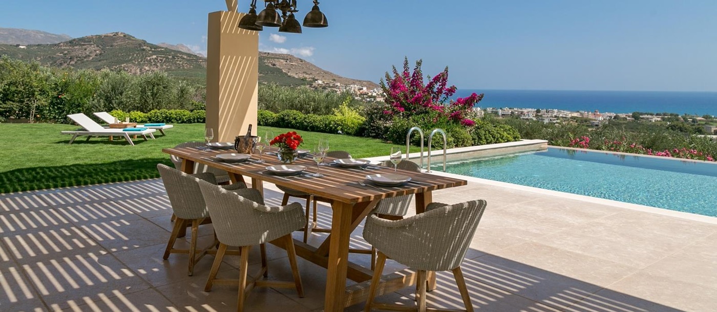 Covered outdoor dining area with table, chairs, hanging lights and pool, garden and sea view at Villa Ianira on Crete, Greece