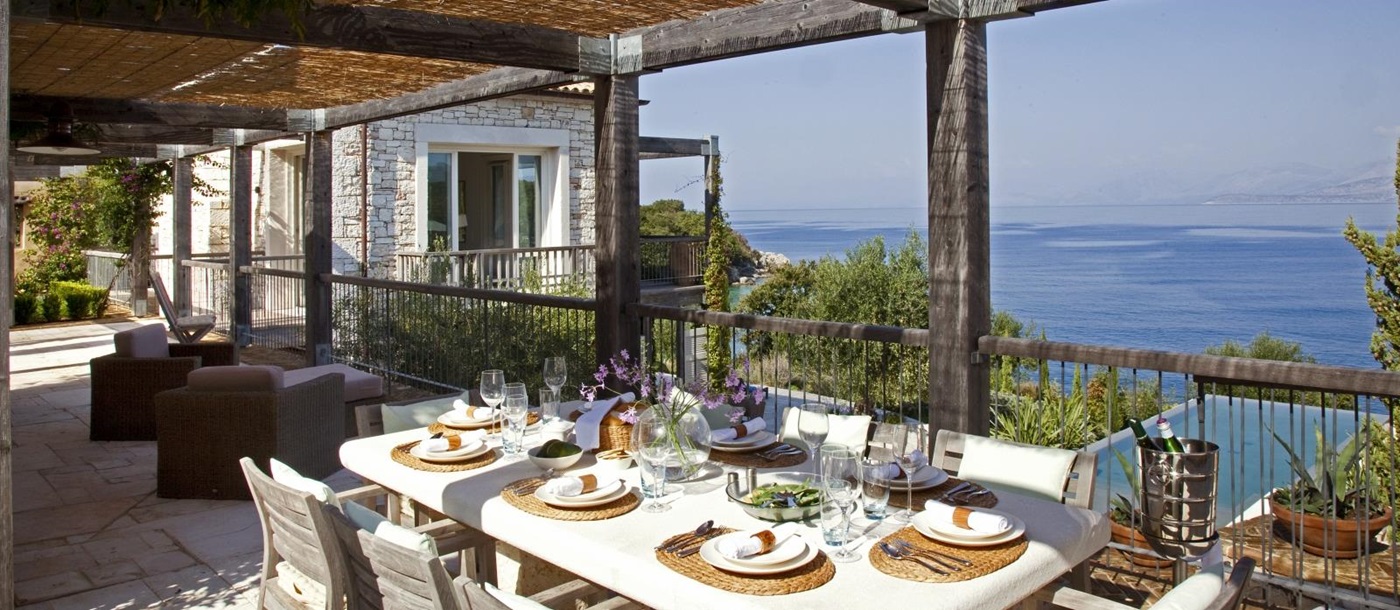 Covered outdoor dining area on balcony with table, chairs, armchairs and sea view at Villa Icarius on Corfu, Greece