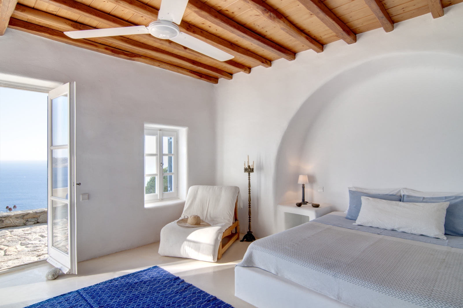 Bedroom with double bed, bedside table, lamp, armchair, ceiling fan and access to terrace at Villa Mari on Mykonos, Greece