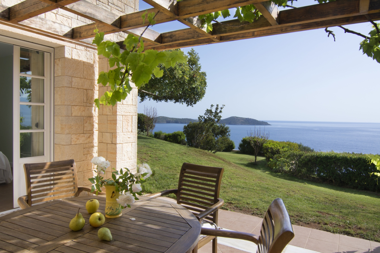 Outdoor dining area with table, chairs, flowers, fruit, arbour, vine and sea view at Villa Selene on Crete, Greece