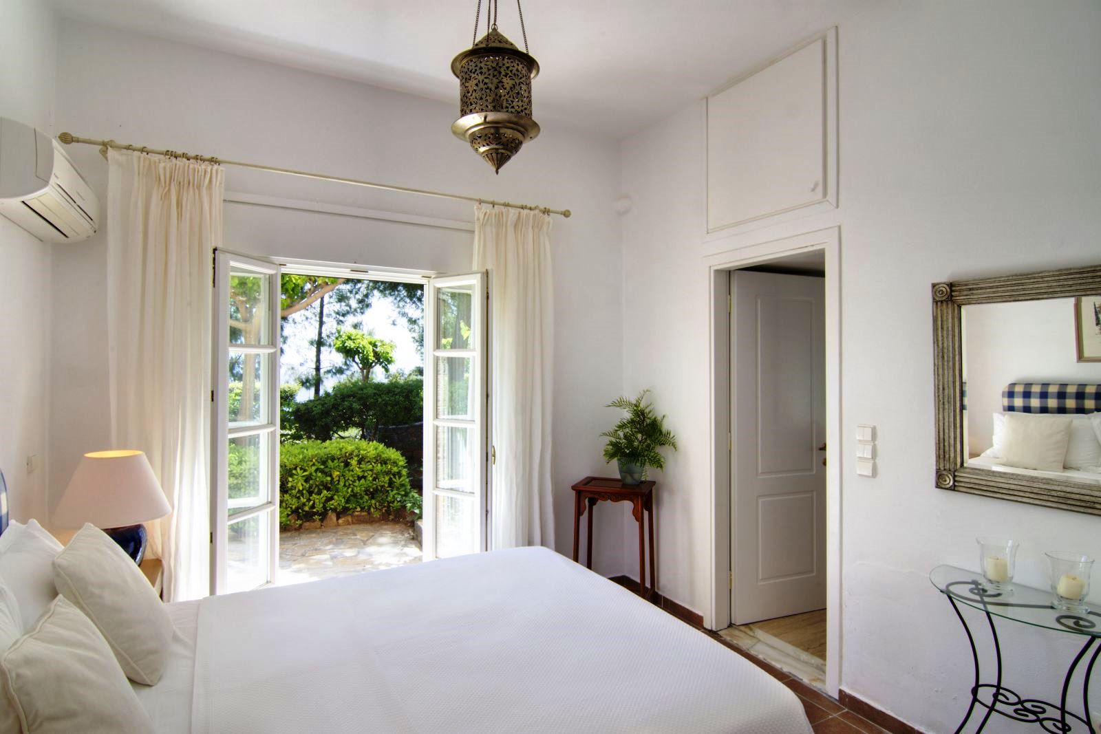 White double bedroom with open glass doors out onto the garden area and white wooden door into an ensuite bathroom at Villa Selene in Crete