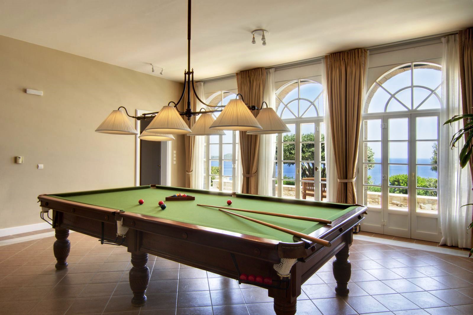 Billiard table with overhanging light fixture on a brown tiled floor in a room with floor to ceiling glass doors that look out over the sea at Villa Selene in Crete