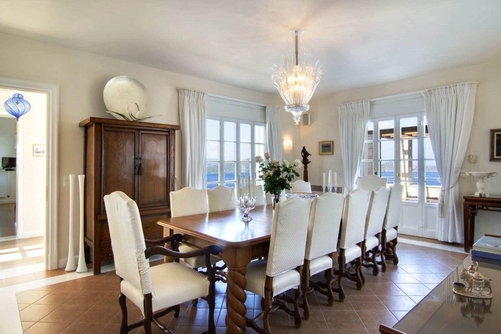 Dining room with brown tiled floor and long wooden table with twelve white cushioned chairs at Villa Selene in Crete