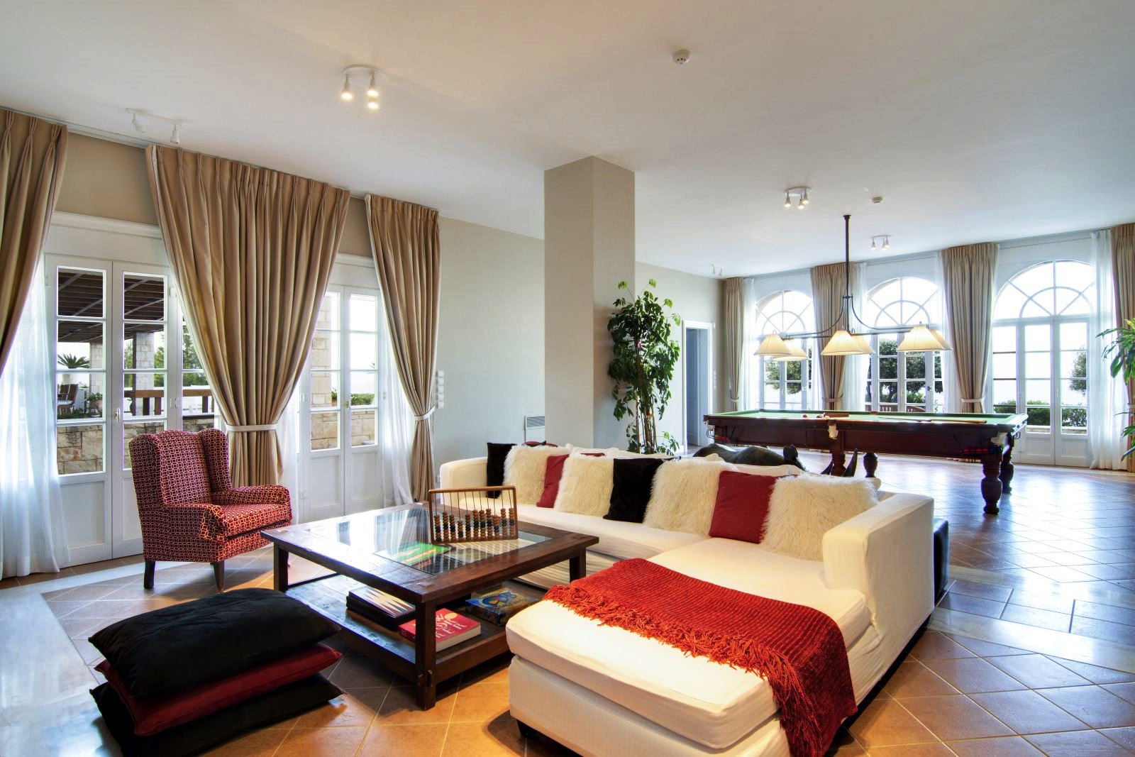 Seating area with large white corner sofa with red, white and black cushions and large coffee table with billiard table behind at Villa Selene in Crete