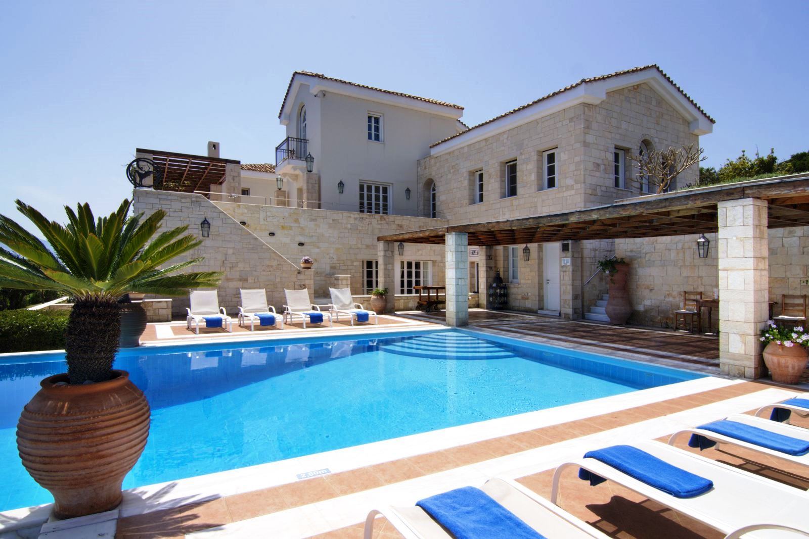 View over the pool that has white sun loungers with blue towels on them with Villa Selene in Crete in the background