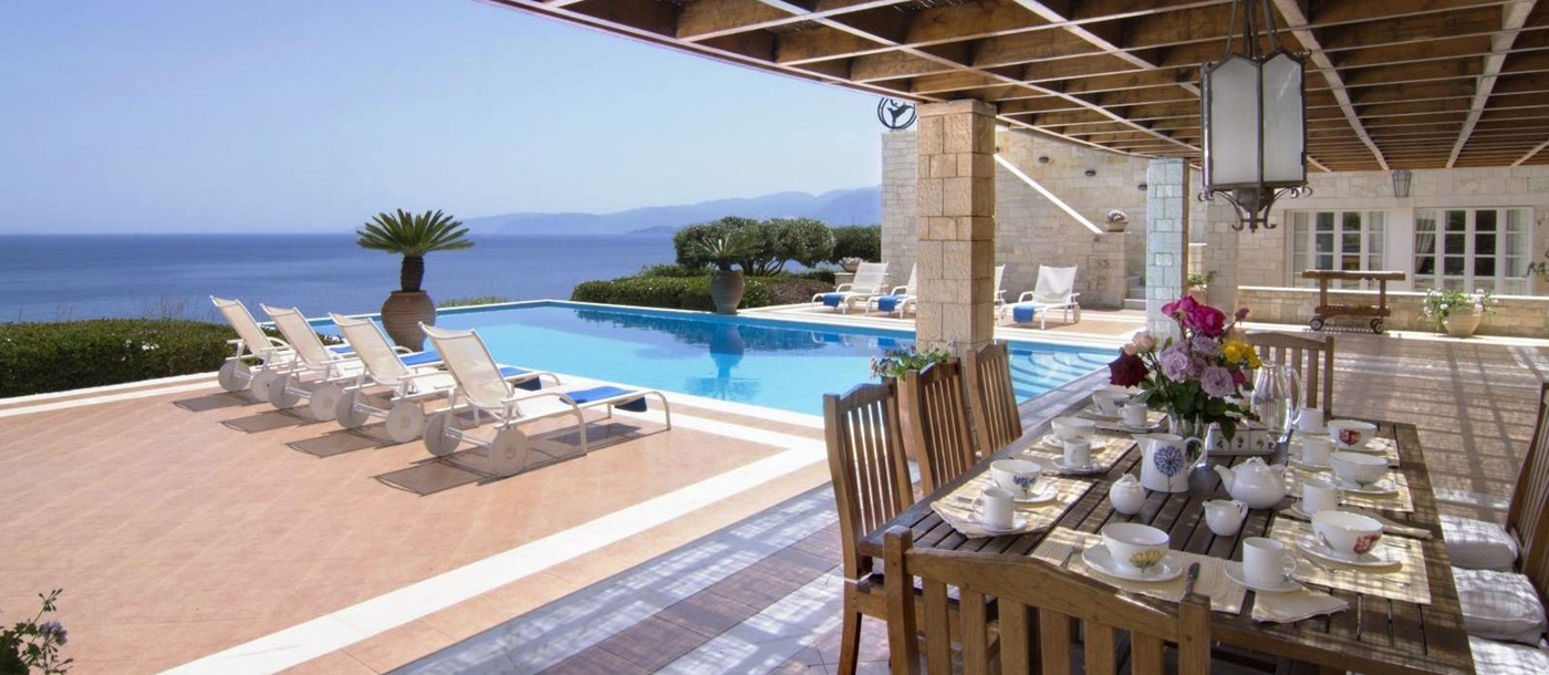Covered terrace area with wooden table set for breakfast and chairs by the side of the pool at Villa Selene in Crete